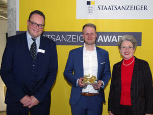 2 Männer und eine Frau. Der Mann in der Mitte hält die Trophäe in der Hand.