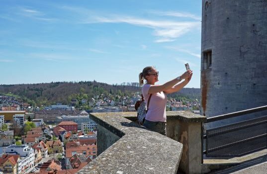 Eine Frau steht an einer Brüstung und macht ein Selfie. Hintergrund ist eine Stadt zu sehen.