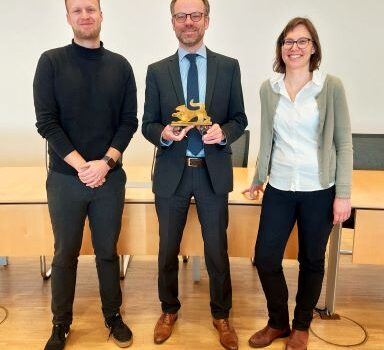 2 Männer und eine Frau stehen vor einem Besprechungstisch. Der Mann in der Mitte hält die Trophäe