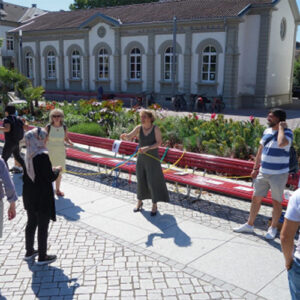 Mehrere Menschen stehen im lockeren Kreisauf einem Platz vor roten Bänken. Im Hintergrund ist eine Hausfassade zu sehen.