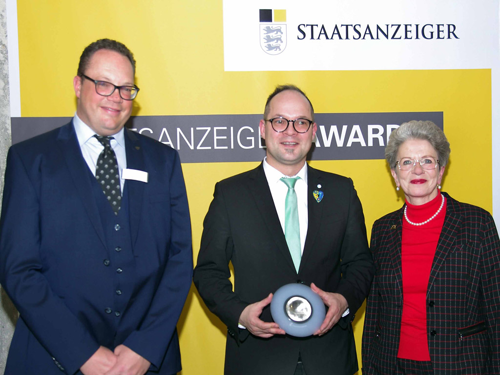 2 Männer und eine Frau stehen vor dem gelben Rollup. Der Mann in der Mitte hält die Trophäe