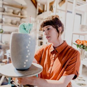 Die Töpferin Lyn Löschel bemalt in einem Atelier mit einem feinen Pinsel eine große, graue Keramikvase. Sie trägt ein orangefarbenes Hemd und konzentriert sich auf ihre handwerkliche Arbeit.