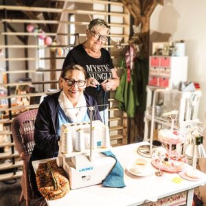 Zwei Frauen in einer kreativ eingerichteten Werkstatt, eine sitzt an einer Nähmaschine, die andere steht hinter ihr. Sie arbeiten gemeinsam an einem Textilprojekt, umgeben von Nähzubehör und Dekoration.