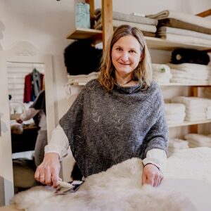 Eine Frau steht in einem Laden mit Woll- und Fellprodukten und bearbeitet ein Schaffell mit einem Werkzeug. Sie lächelt und trägt einen grauen Poncho über einer weißen Bluse.