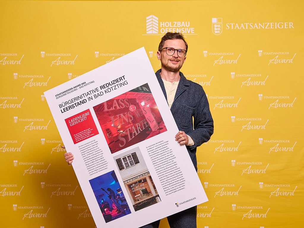 2. Platzierte der Kategorie Bürger:innenbeteiligung steht vor einer Pressewand und hält eine Projektcollage in die Kamera.