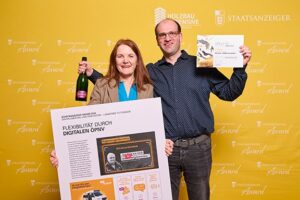 Ein Mann und eine Frau stehen vor einer Pressewand und halten Urkunde und Projekt Collage in die Kamera.