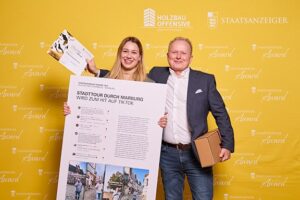 Zwei Personen lächeln vor einer gelben Pressewand mit Logos und halten eine Auszeichnung sowie ein Plakat mit dem Titel „Stadttour durch Marburg wird zum Hit auf TikTok“.