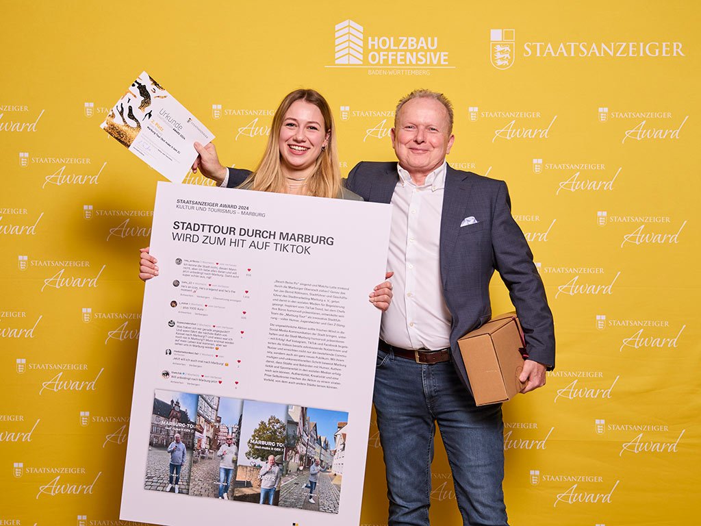 Zwei Personen lächeln vor einer gelben Pressewand mit Logos und halten eine Auszeichnung sowie ein Plakat mit dem Titel „Stadttour durch Marburg wird zum Hit auf TikTok“.
