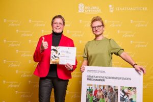 Zwei Frauen stehen glücklich vor einer Pressewand und halten Urkunde und Projekt Collage in die Kamera.