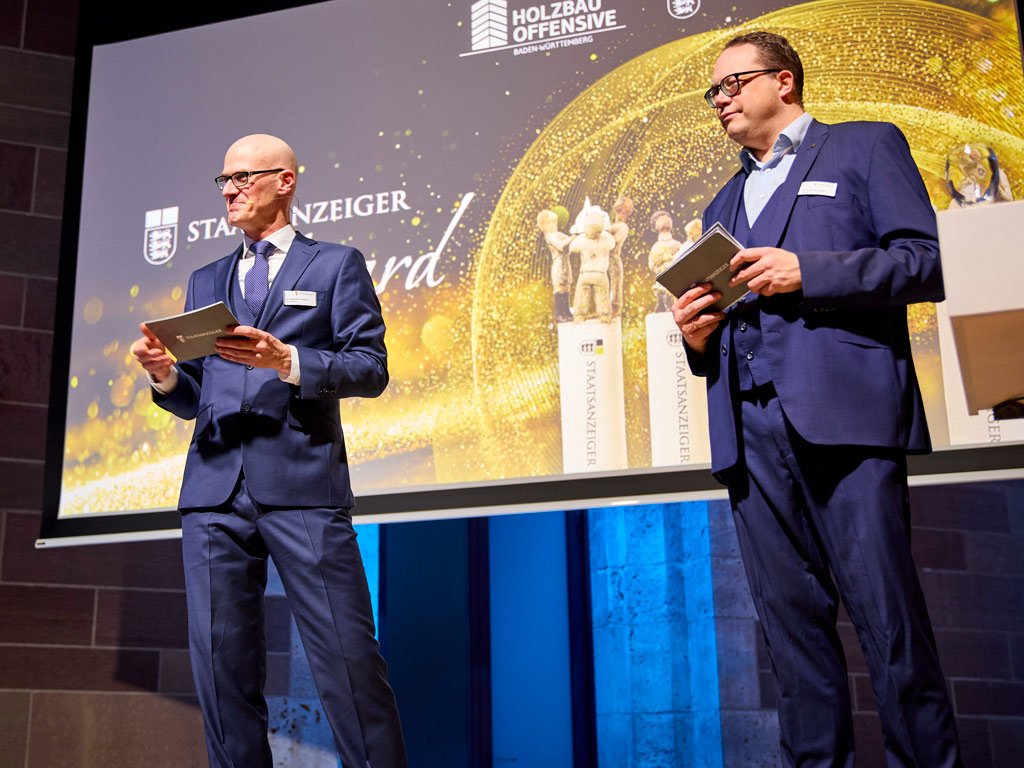 Geschäftsführer Dr. Alexander Teutsch und Chefredakteur Dr. Rafael Binkowski stehen vor einer großen Leinwand auf der das Logo des Awards abgebildet ist. Vor ihnen steht ein Stehpult mit Mikrofon