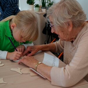Ein älterer Mensch und ein Kind basteln zusammen an einem Tisch. Das Kind zeichnet konzentriert auf ein Stück Papier, während die Seniorin assistiert.