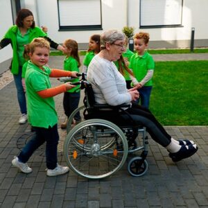 Ein Junge in einem grünen T-Shirt schiebt eine ältere Dame im Rollstuhl über einen gepflasterten Weg, begleitet von anderen Kindern.