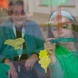Ein älterer Mensch und ein Kind malen gemeinsam auf eine Glasscheibe. Das Kind trägt ein grünes T-Shirt und konzentriert sich auf das Malen.