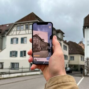 Eine Hand hält ein Smartphone, das ein altes, historisches Bild eines Straßenzugs zeigt. Im Hintergrund ist derselbe Ort in der Gegenwart zu sehen.