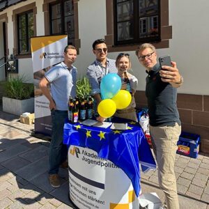 Vier Personen machen ein Selfie hinter einem Tisch mit Europaflagge und gelb-blauen Luftballons. Im Hintergrund sind Banner der "Akademie Jugendpolitik e.V." zu sehen.