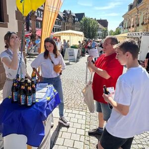Eine Frau erklärt etwas an einem mit einer Europaflagge bedeckten Tisch mit Bierflaschen. Zwei junge Männer und eine Frau hören aufmerksam auf einem sonnigen Marktplatz zu.