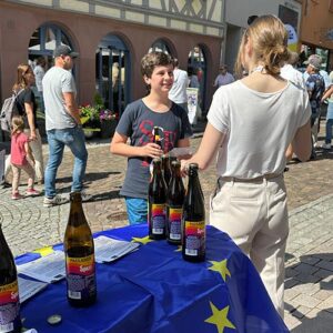 Ein Junge steht an einem Tisch mit einer Europaflagge, auf dem Bierflaschen stehen. Eine Frau im weißen Oberteil spricht mit ihm auf einem belebten Platz.