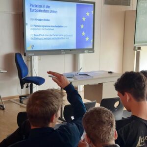 Jugendliche sitzen in einem Klassenraum und heben die Hände. Auf dem Bildschirm vorne ist eine Präsentation zum Thema "Parteien in der Europäischen Union" zu sehen.