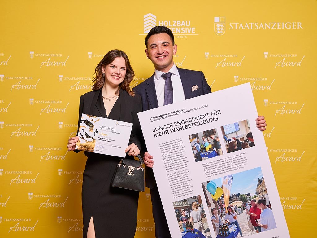Eine junge Frau und ein Mann posieren vor einer gelben Wand mit dem Logo des "Staatsanzeiger Award". Die Frau hält eine Urkunde, der Mann ein großes Poster mit dem Titel "Junges Engagement für mehr Wahlbeteiligung"