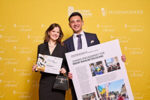 Eine junge Frau und ein Mann posieren vor einer gelben Wand mit dem Logo des "Staatsanzeiger Award". Die Frau hält eine Urkunde, der Mann ein großes Poster mit dem Titel "Junges Engagement für mehr Wahlbeteiligung"