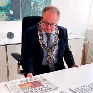 Bürgermeister Bernd Hornikel in einem Anzug mit Amtskette sitzt an seinem Schreibtisch und wirft einen kurzen Blick auf die Schorndorfer Zeitung. Im Hintergrund ist ein minimalistisches Büro mit Dekoration zu erkennen.