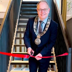 Bürgermeister Bernd Hornikel in einem Anzug mit Amtskette schneidet mit einer Schere ein rotes Band durch, während er in die Kamera lächelt. Im Hintergrund ist eine Treppe zu sehen.