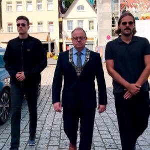 rei Männer laufen nebeneinander über einen gepflasterten Platz. In der Mitte trägt ein Mann einen dunklen Anzug und eine Amtskette, begleitet von zwei Männern in legerer Kleidung und Sonnenbrillen.