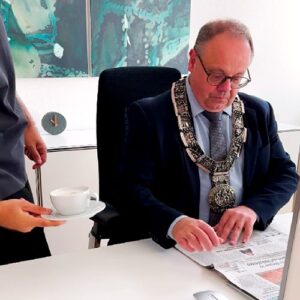 Bürgermeister Bernd Hornikel in einem Anzug mit Amtskette sitzt an seinem Schreibtisch und liest die Schorndorfer Zeitung. Sein Assistent stellt ihm eine Tasse Kaffee hin.
