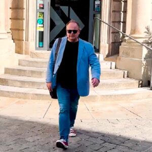 Ein Mann mit Sonnenbrille, blauem Sakko und schwarzem Shirt geht entschlossen eine Treppe hinunter. Er trägt eine Umhängetasche und Sneakers.
