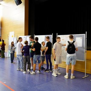 Eine Gruppe von Jungen und Mädchen diskutiert vor Stellwänden mit Plakaten in einer Sporthalle. Im Hintergrund ist eine Bühne erkennbar.