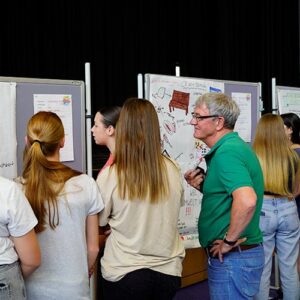 Jugendliche stehen vor Stellwänden und betrachten Plakate mit Vorschlägen. Ein älterer Mann im grünen Poloshirt hört aufmerksam zu.