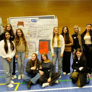 Eine Gruppe von Schülerinnen steht vor einer Stellwand und präsentiert ihre Arbeit. Auf der Wand sind Plakate und Zeichnungen zu sehen, die kreative Vorschläge darstellen.