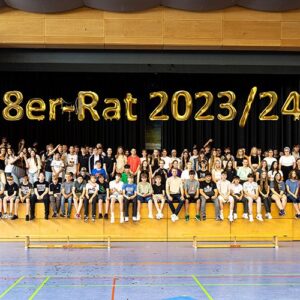 Eine große Gruppe von Schülerinnen und Schülern sitzt auf einer Tribüne in einer Sporthalle. Über ihnen hängen goldene Luftballons mit der Aufschrift „8er-Rat 2023/24“