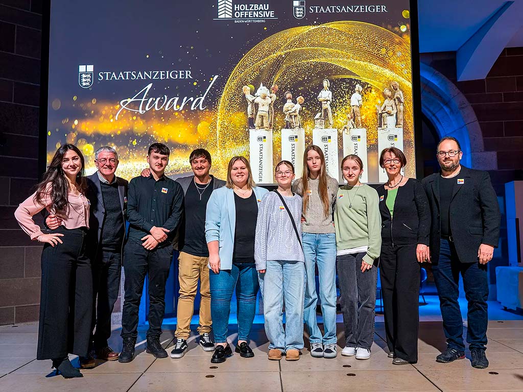 Eine Gruppe von Personen steht auf einer Bühne vor einer Projektion mit dem Titel „Staatsanzeiger Award“. Im Hintergrund sind goldene Trophäen abgebildet. Die Personen lächeln und posieren für ein Gruppenfoto.