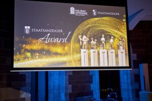 Bühne mit einer großen Leinwand, auf der „Staatsanzeiger Award“ und Trophäen vor einem funkelnden Hintergrund zu sehen sind.
