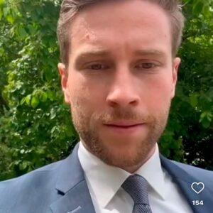 Ein Mann im Anzug steht im Freien vor einer grünen, baumreichen Kulisse. Er blickt direkt in die Kamera.