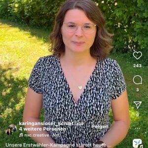 Eine Frau mit kurzen Haaren und einer Brille steht vor einer grünen Wiese. Sie trägt ein gemustertes Kleid und spricht in die Kamera.