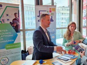 Ein Mann im Anzug und eine Frau im karierten Blazer stehen an einem Messestand. Sie präsentieren Materialien und sprechen mit einer weiteren Person. Im Hintergrund sind Banner mit Informationen zur Weiterbildungsplattform für Kommunen zu sehen. Der Raum ist hell und modern gestaltet.