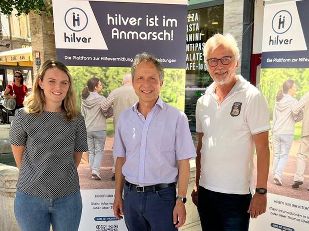 Drei Personen stehen vor zwei Roll-Ups mit der Aufschrift „Hilver ist im Anmarsch!“ und lächeln in die Kamera. Im Vordergrund ein Tisch mit Flyern zum Projekt.