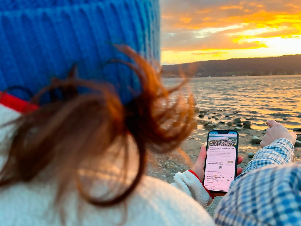 Eine Person mit blauer Mütze und wehendem Haar hält ein Smartphone mit einer geöffneten App in der Hand. Im Hintergrund sind ein See, eine untergehende Sonne und eine hügelige Landschaft zu sehen.