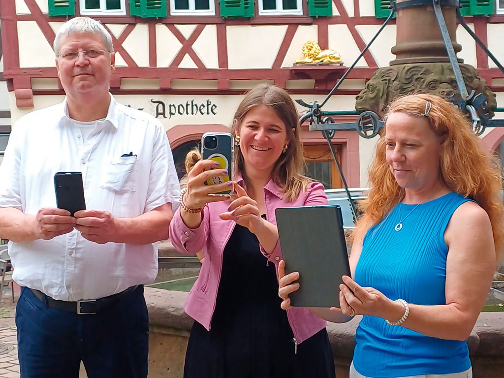 Drei Personen stehen vor einem historischen Fachwerkgebäude und halten Smartphones oder Tablets in den Händen. Eine Frau in der Mitte lächelt in die Kamera, während sie ein Handy hält.