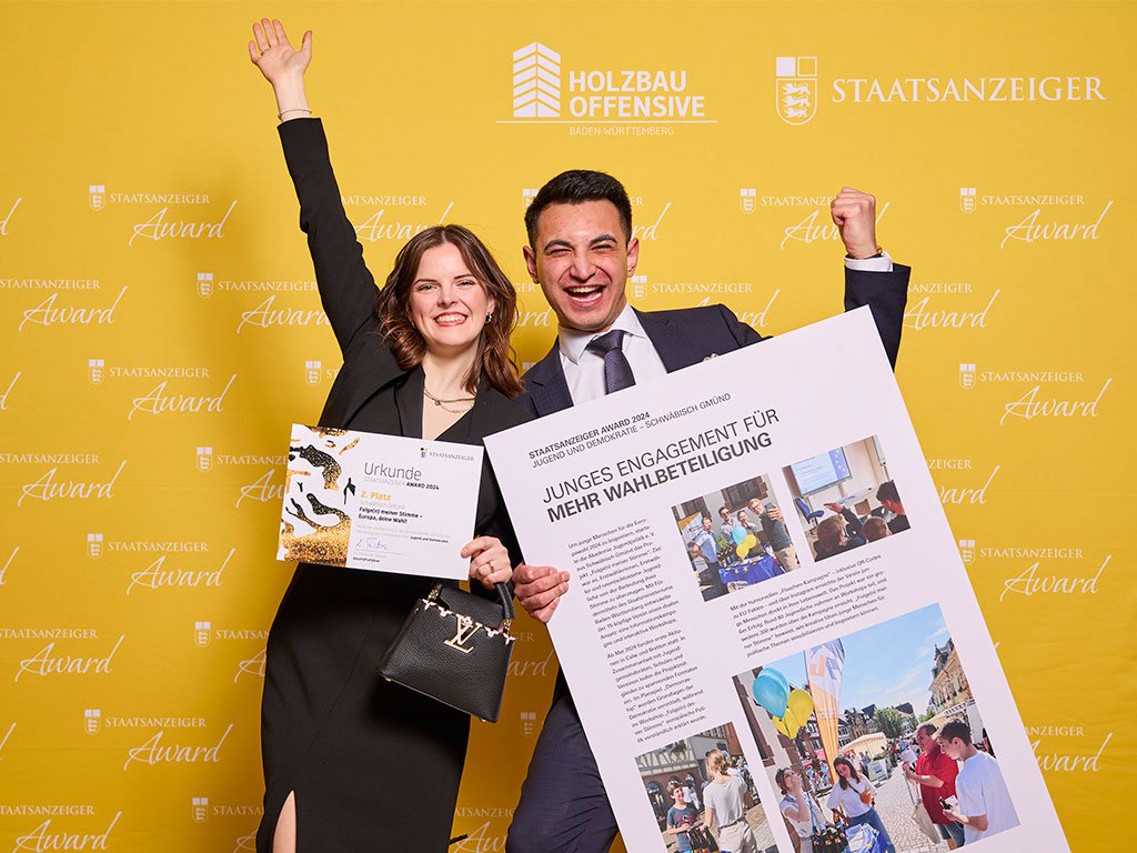 Eine junge Frau und ein Mann posieren vor einer gelben Wand mit dem Logo des "Staatsanzeiger Award". Die Frau hält eine Urkunde, der Mann ein großes Poster mit dem Titel "Junges Engagement für mehr Wahlbeteiligung"
