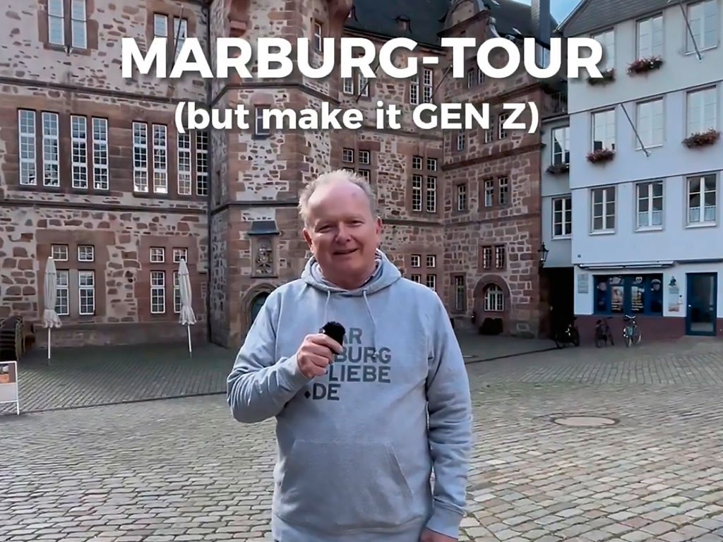 Mann steht auf einem historischen Platz in Marburg mit Mikrofon, Text: „MARBURG-TOUR (but make it GEN Z)“.