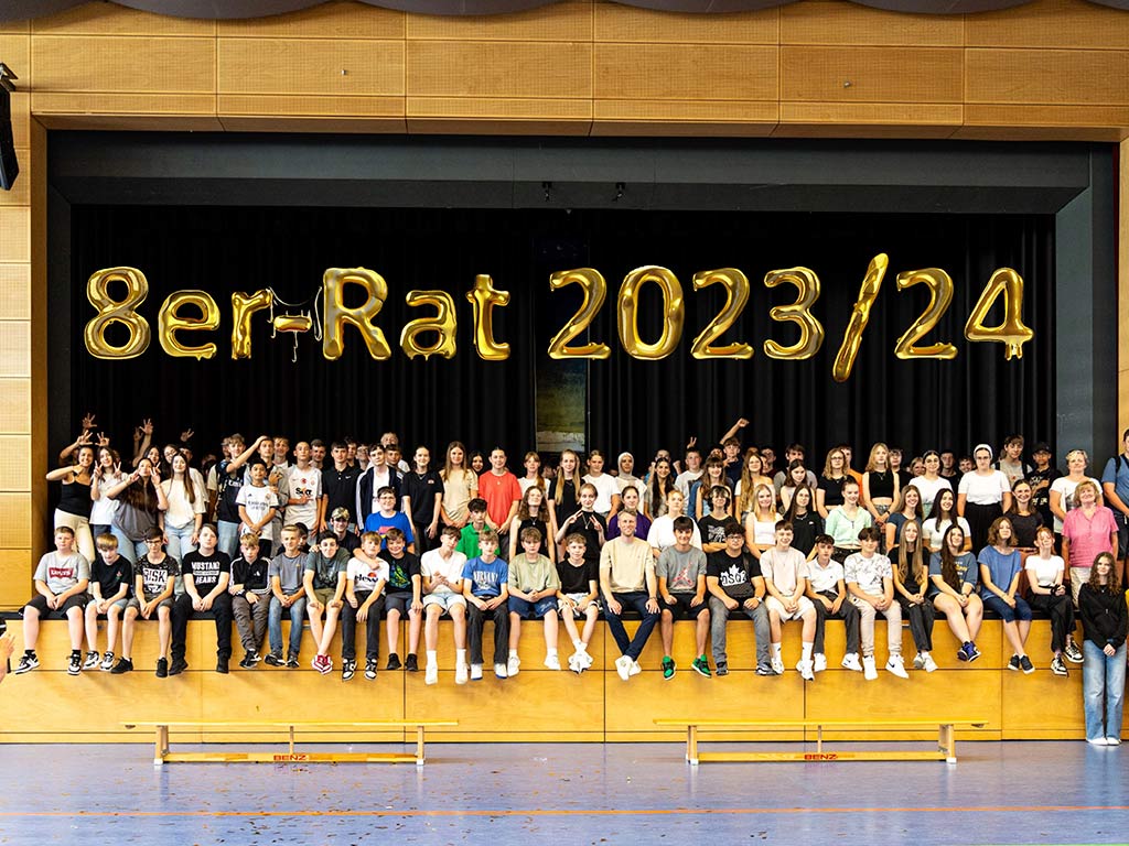 Eine große Gruppe von Schülerinnen und Schülern sitzt auf einer Tribüne in einer Sporthalle. Über ihnen hängen goldene Luftballons mit der Aufschrift „8er-Rat 2023/24“
