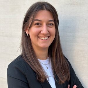 Das Bild zeigt eine Frau mit langen, glatten Haaren, die lächelnd in die Kamera blickt. Sie trägt ein schwarzes Sakko über einem weißen Oberteil und steht vor einer hellen, strukturierten Wand.