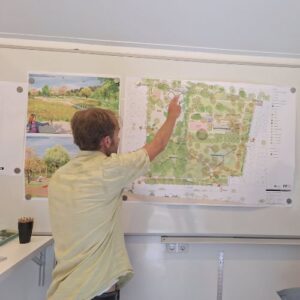 Eine Person zeigt auf einen großen Bauplan an einer Wand; im Raum sind weitere Pläne/Karten an Pinnwänden oder Wänden zu sehen.