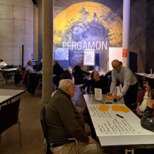 Veranstaltungssaal mit mehreren Teilnehmenden an Tischen; im Hintergrund ist eine große Projektion mit der Aufschrift „PERGAMON“ sichtbar.