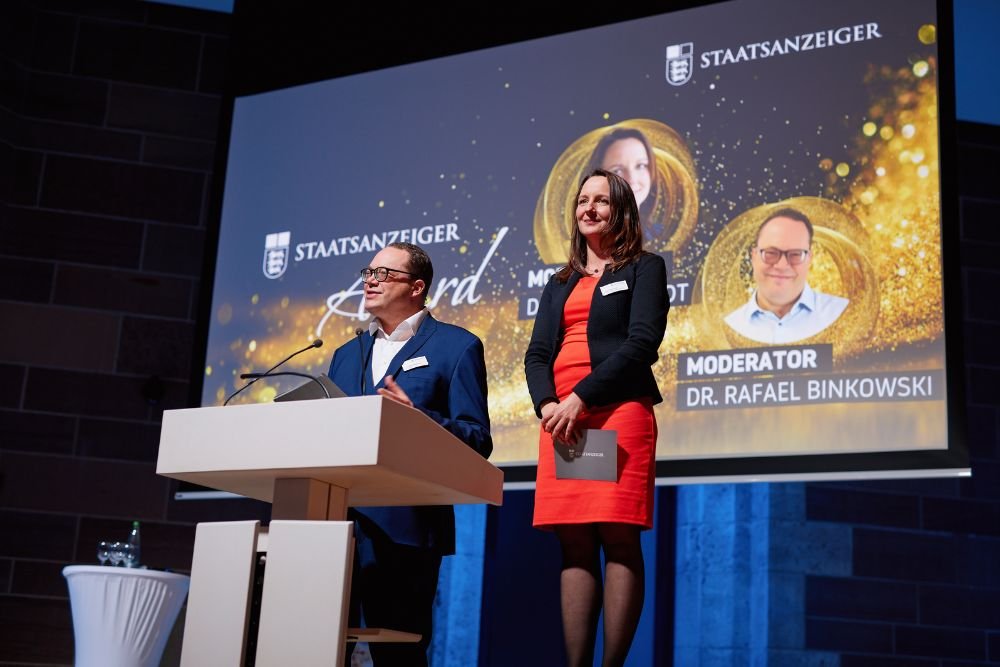 Dana Reinhardt und Rafael Binkowski stehen auf der Bühne am Rednerpult bei der Verleihung des Staatsanzeiger Awards 2025 vor einer Präsentationsfolie.