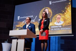 Staatsanzeiger Award 2025 Dana Reinhardt und Rafael Binkowski stehen auf der Bühne am Rednerpult bei der Verleihung des Staatsanzeiger Awards 2025 vor einer Präsentationsfolie.
