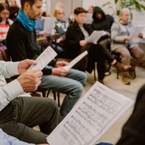 Publikum sitzt in einem Raum und verfolgt eine musikalische Aufführung mit ausgeteilten Notenblättern